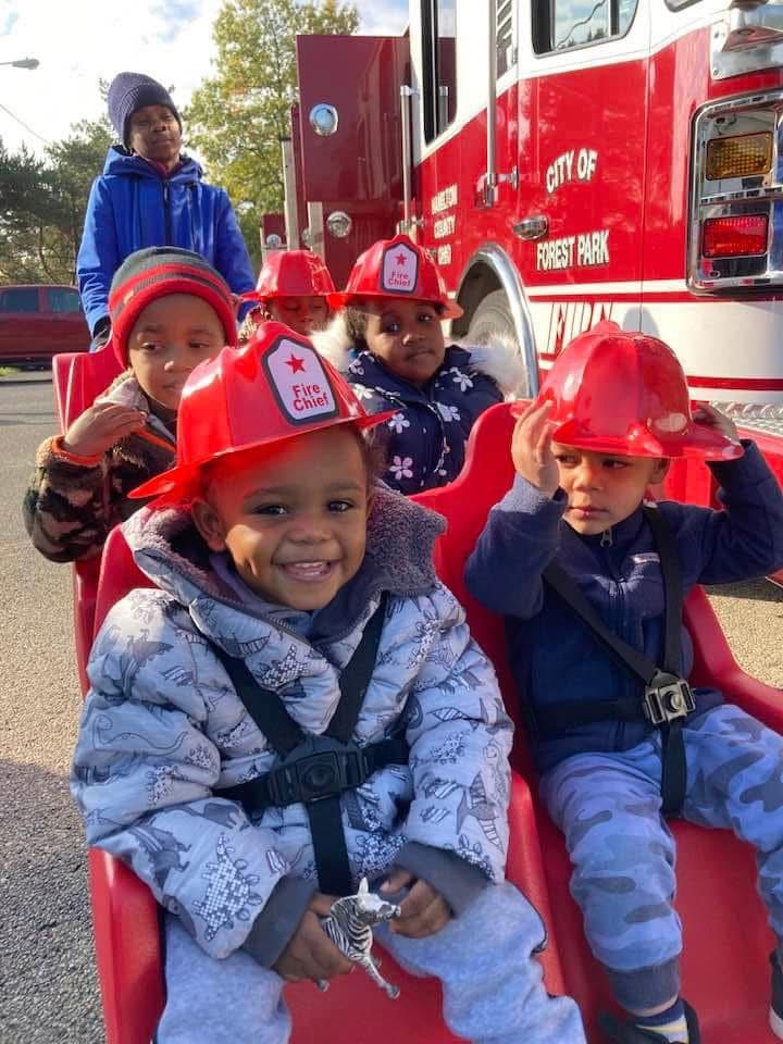 Young children wearing red fire chief hats beside a City of Forest Park fire engine